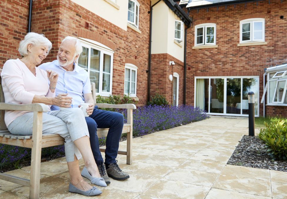 An elderly couple sits on a bench outside of a senior housing facility.