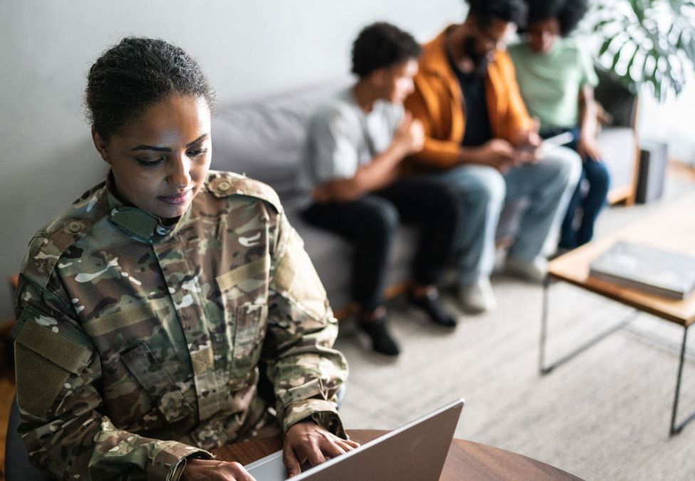 A military servicewoman works at a laptop while her family look sit on a couch.