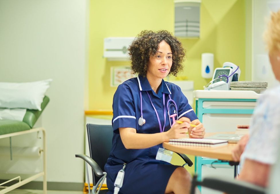 medical professional with dark curly hair sitting at desk talking to patient