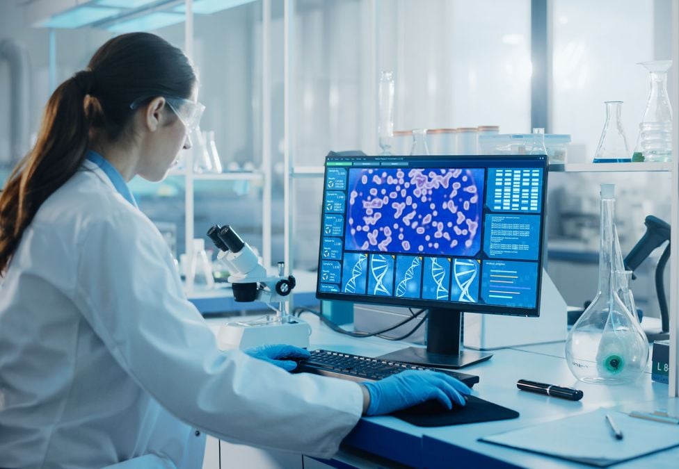 An image of a woman with white skin and brunette ponytail who is a scientist working in a lab. She is using a computer with technical items on the screen and wearing a white lab coat.