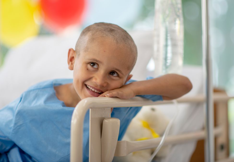 A mixed race oncology patient sits in his hospital bed, in his gown, with his hair shaved. He has an optimistic smile on his face as he receives his Chemotherapy via IV drip treatment.